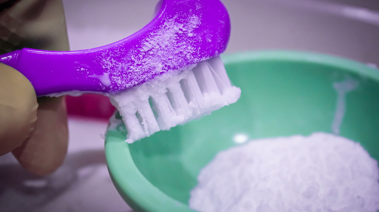 A gloved hand applies a baking soda paste in a small bowl to a soft-bristled brush.