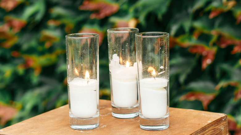 Three white burning candles inside of tall glass hurricane holders, set on a wooden surface against a lush, green background.