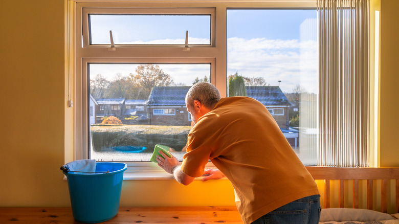 A man cleaning a window with a sponge while a blue bucket sits nearby