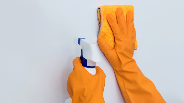 Hands in orange rubber gloves holding a spray bottle and cloth up to a white wall to clean it