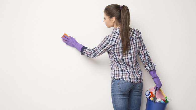 A woman in a flannel, jeans, and purple rubber gloves wipes a wall down with a sponge with one hand and holds a bucket of cleaning supplies in the other