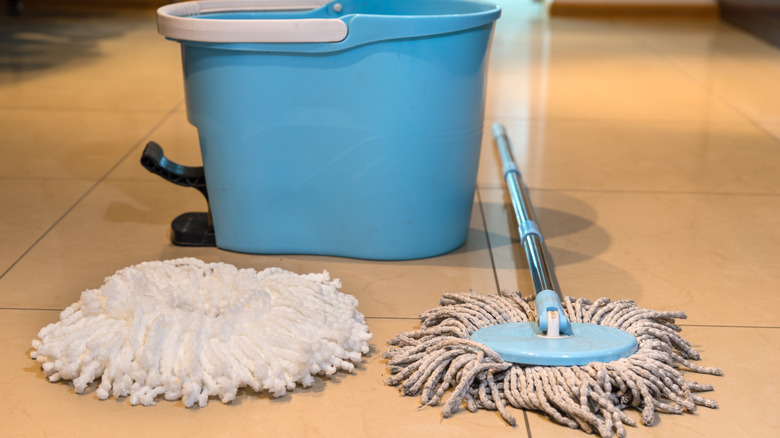 An old dirty mop with a new mop head on a floor with a blue bucket in the background