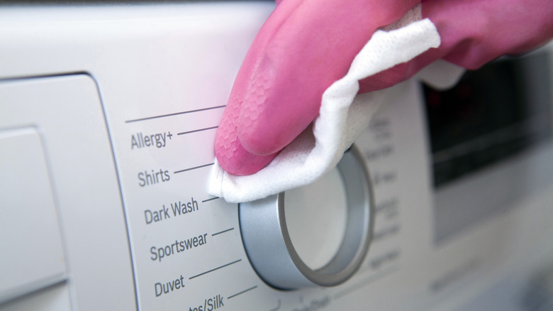 Close-up of a hand in a rubber glove wiping down a washing machine dial with a disinfectant disposable wipe