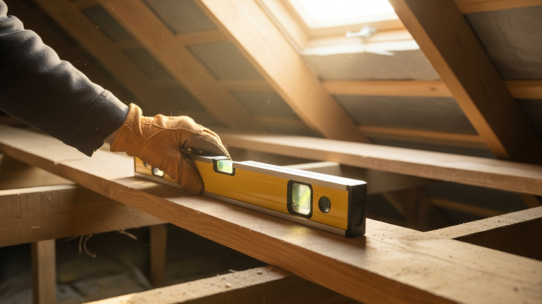 A carpenter uses a level on a beam in an unfinished attic.