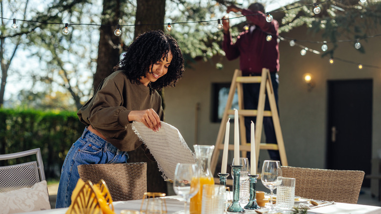One person decorates a patio table while another hangs lights in the background
