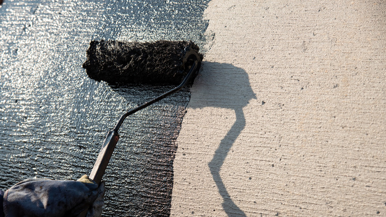 A hand using a roller to apply black paint to concrete