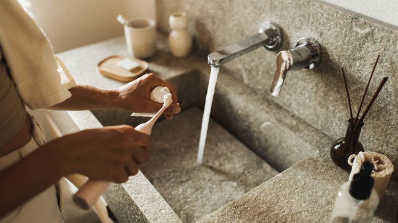 person putting toothpaste on toothbrush with water running in stone sink
