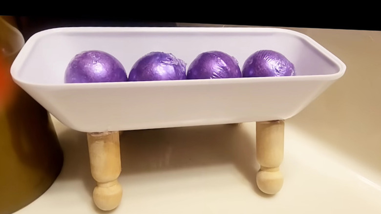 A DIY bathroom tray made from wooden peg dolls and a plastic butter dish with purple bath bombs inside.