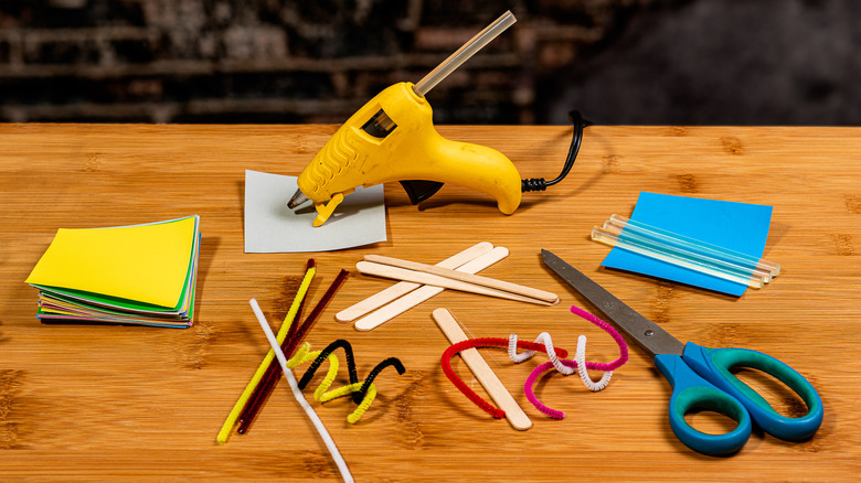 Multiple crafting tools such as glue glue, chenille wires, and ice cream sticks over wooden table.