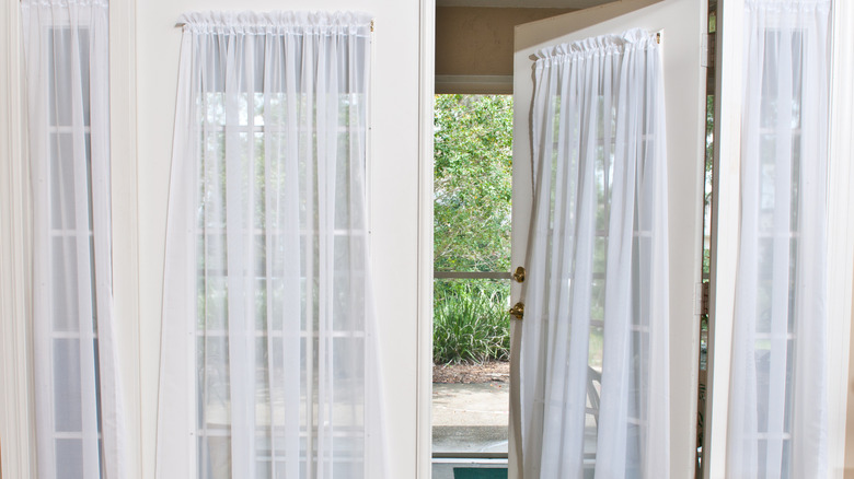 A set of french doors with white, sheer curtains open to a green space outside.