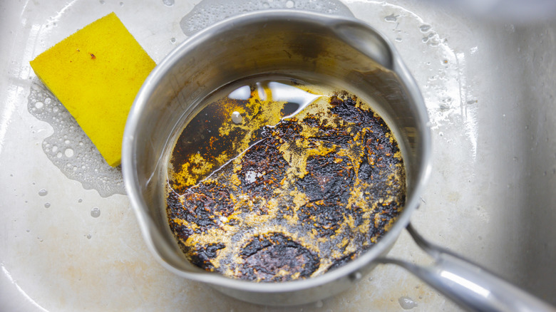 Empty silver pot with charred stains on bottom sitting inside a sink next to sponge.