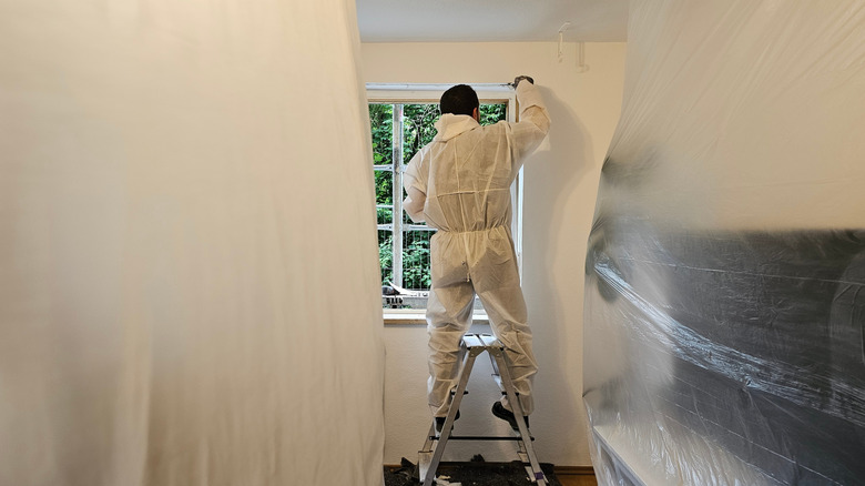 Man on ladder with protective gear and plastic sheeting on furniture for cleaning.