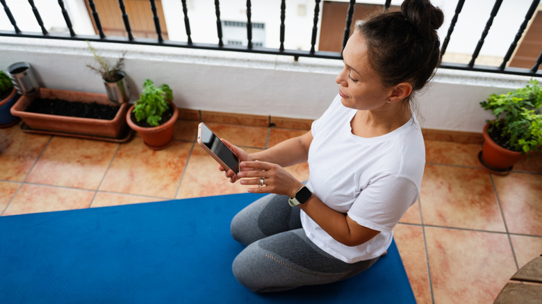 Woman on a yoga mat on a sparsley decorated balcony using her smartphone