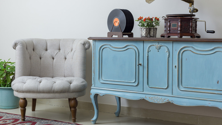 Vintage interior of retro off-white armchair, vintage wooden light blue sideboard, old phonograph (gramophone), and vinyl records on background of beige wall, tiled porcelain floor, and red carpet.