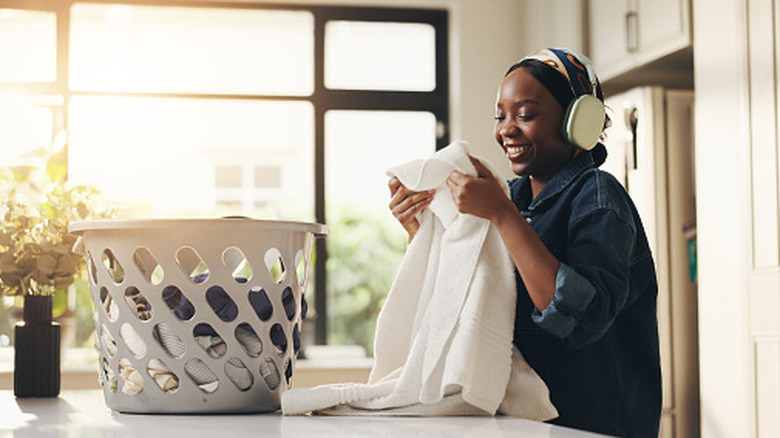 A woman holding up a shift from a basket of laundry while wearing over-ear headphones and smiling