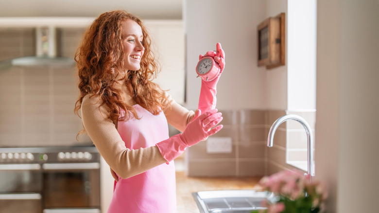 A woman in pink gloves and apron looking at a pink timer while smiling in a bright kitchen