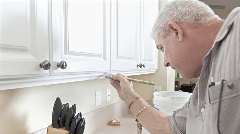 A person paints a wooden kitchen cabinet with white paint.