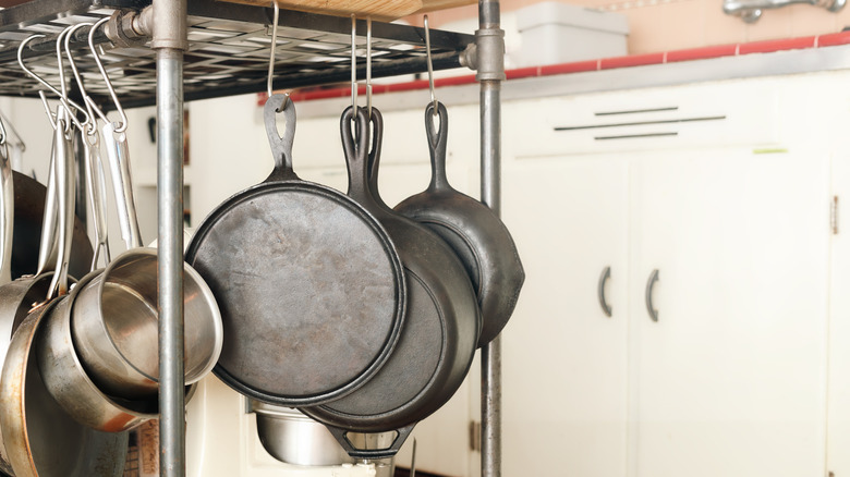A bunch of cast iron and steel pans hanging from a metal shelf on hooks in a kitchen.