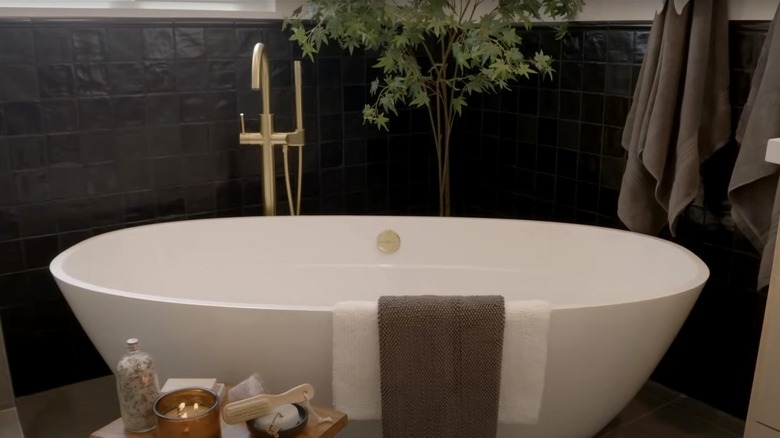 A white soaking tub with gold hardware sits in front of a black tile wall