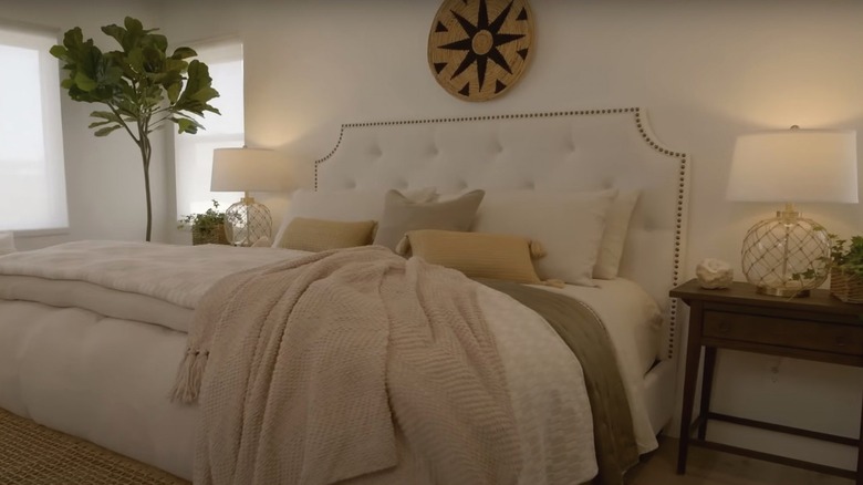 A large woven tray and coastal glass lamps surround a large bed piled high with neutral blankets and pillows