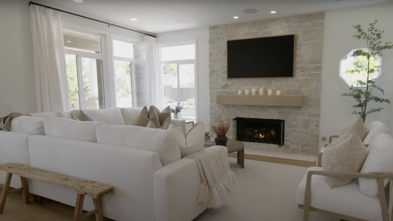 A large stone fireplace is surrounded by large windows dressed with white curtains and a white sectional sofa