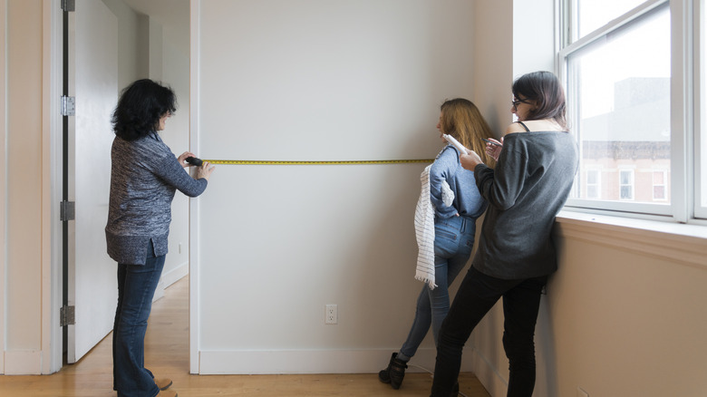 Three women measuring a wall in an empty room and taking notes