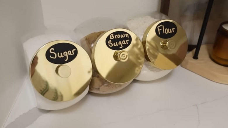 Three candy jars with gold lids hold sugar, brown sugar, and flour on a high-end countertop.