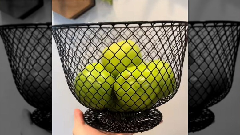 A black wire pedestal fruit bowl holding green apples.