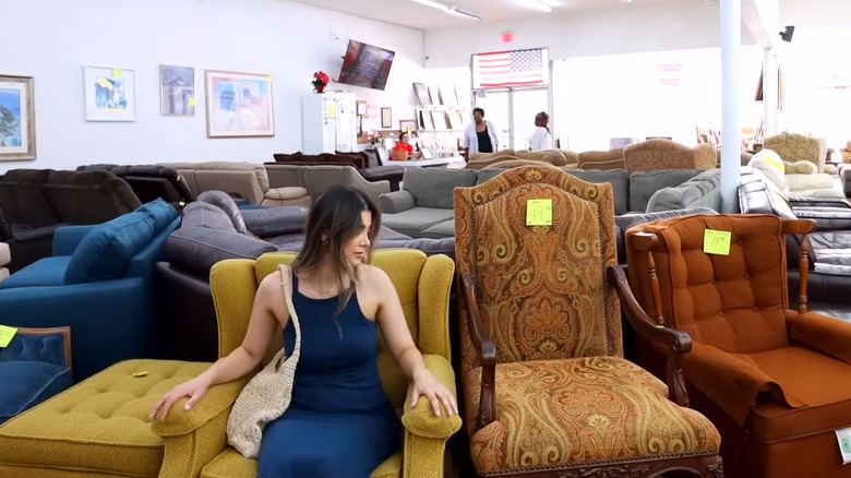 Woman testing out a mustard yellow vintage chair in a thrift store with other vintage chairs next to her