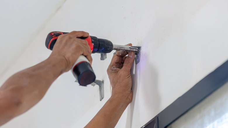 A man's hands using a drill to attach curtain rod brackets into a white wall.