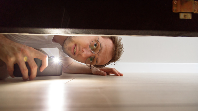 Man looking under the bed with a cell phone flashlight.