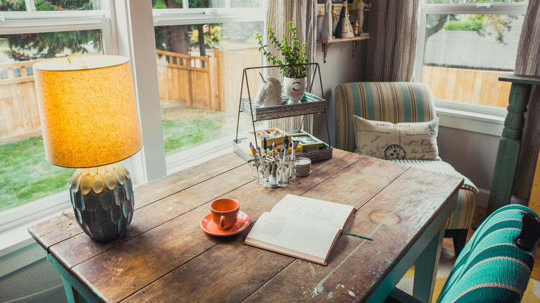 A home office incorporates the cubicle chic decor trend with a rustic farmhouse table, stylish knicknacks, comfy chair, and cute lamp