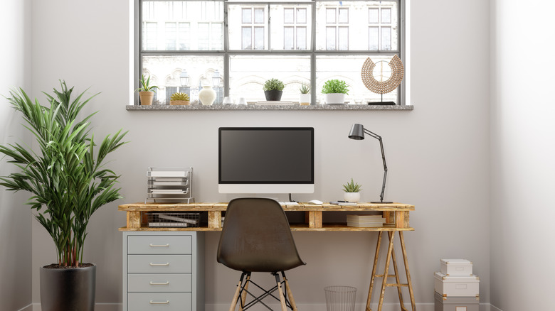 A home office set up in front of a window with small plants on the windowsill and a large one next to the desk