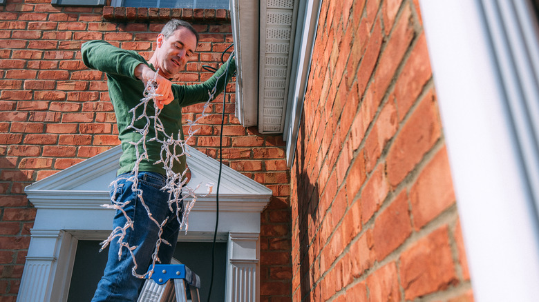 Man on a ladder trying to hang Christmas lights on his home's exterior