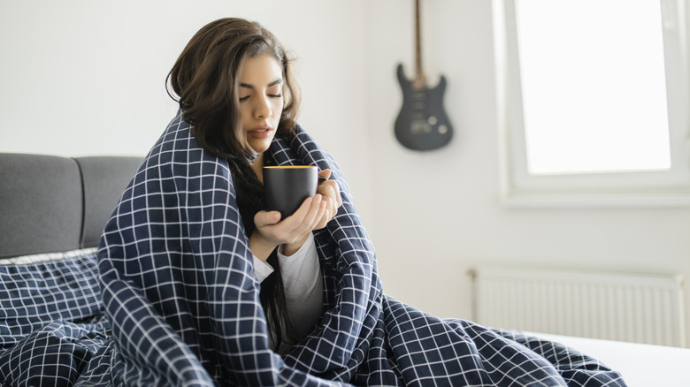 Woman with blanket around herself in bed and holding a mug
