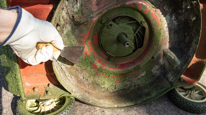 A person using a scraper to clean debris off the rusted deck of a lawn mower