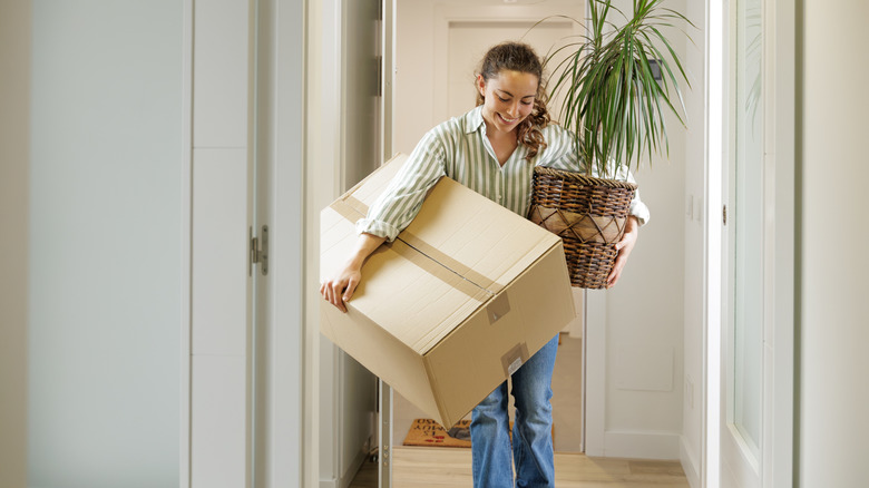 A woman holding a cardboard box and plant in hallway.