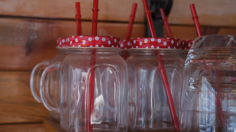 Empty Mason jars with handles, red polka dot lids, and red straws against a wooden wall.
