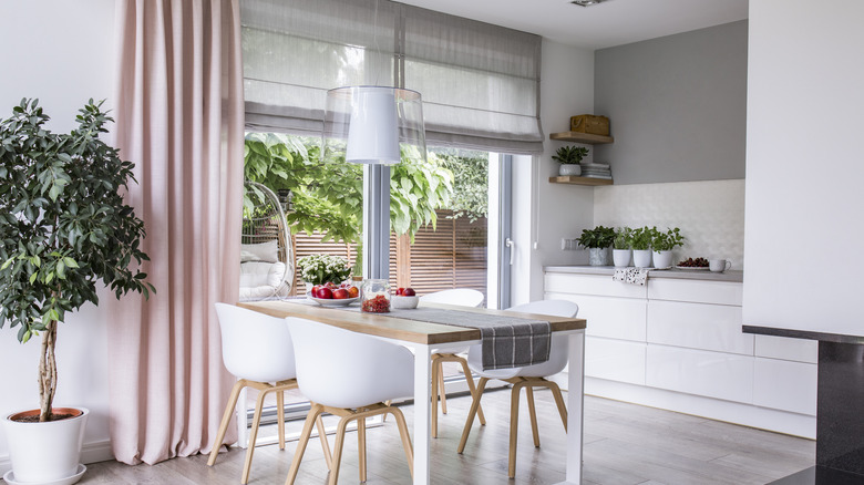 White kitchen table with wooden chairs and a sliding glass door with Roman blinds and pink drape.
