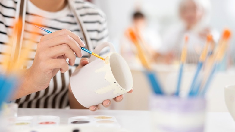 Person painting white coffee mug surrounded by painting supplies
