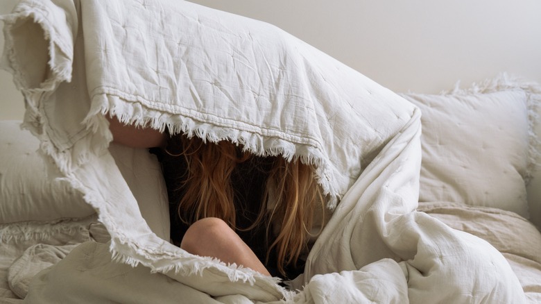 Woman on top of her bed, cocooned in a large blanket, covering her face