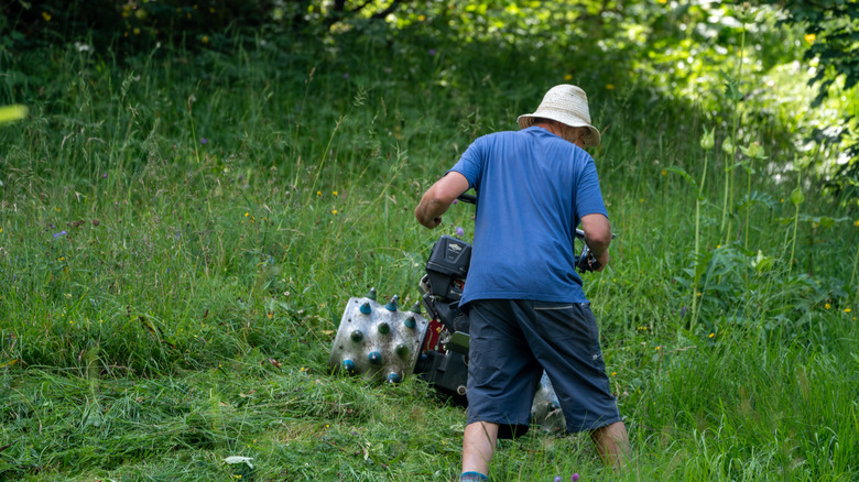 Man in blue shirt and white hat operating a walk-behind mower in tall grass