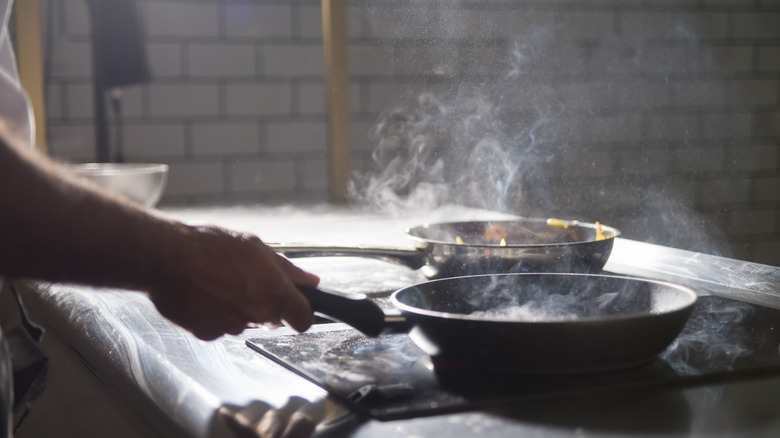 A hand holding a smoking frying pan on a cooktop in a dark room.