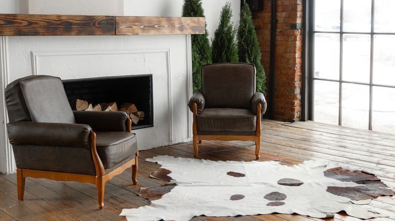 Two brown leather chairs sit in front of a fireplace mantel, with a white and brown cow hide rug in front.