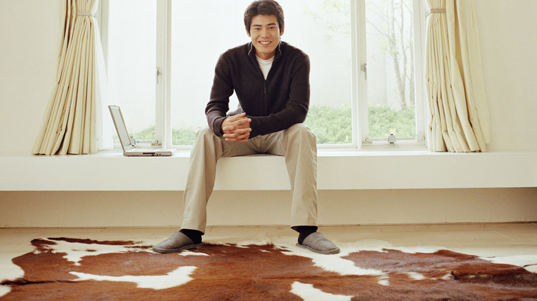A man sits in front of a window with a cow hide rug laid on the floor.
