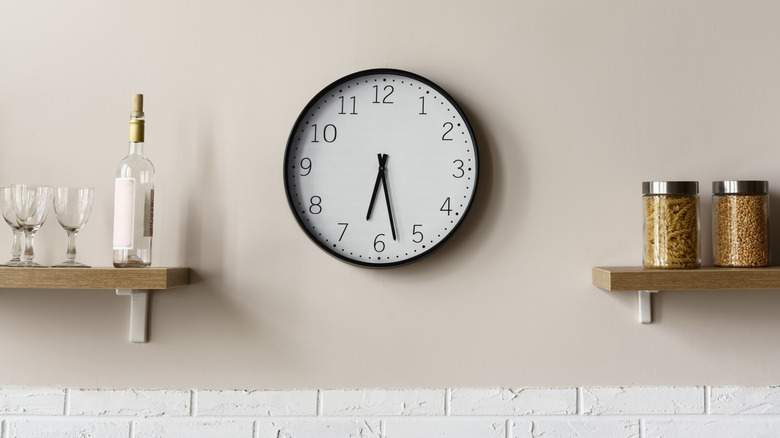 A black and white clock on a kitchen wall surrounded by shelves