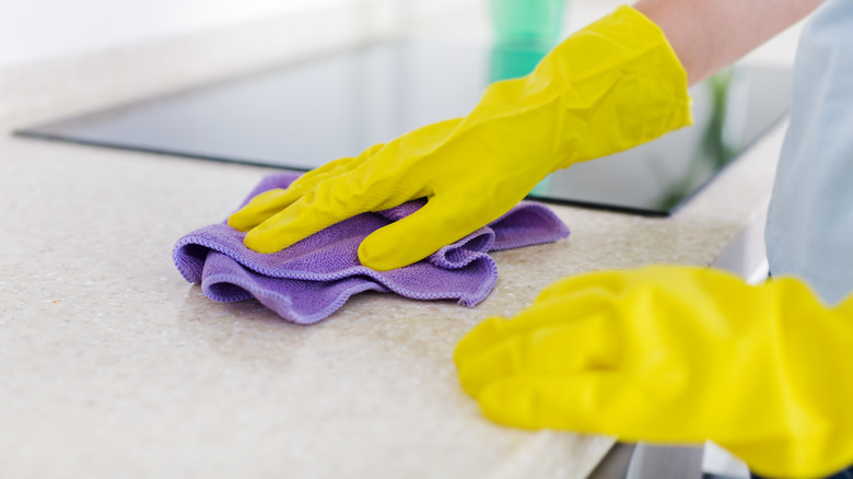 A person wearing yellow rubber gloves wiping a granite countertop with a purple soft cloth
