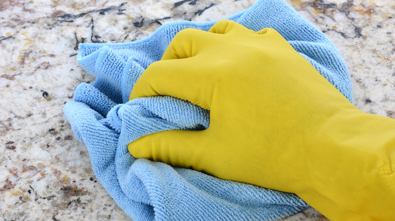 A person wearing yellow rubber gloves wiping granite countertops with a blue microfiber cloth