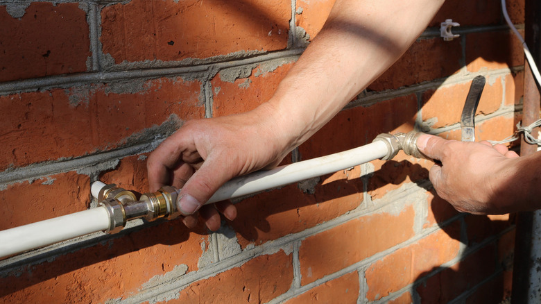 A person installs white water pipes and metal fittings along the outside of a brick walled house.