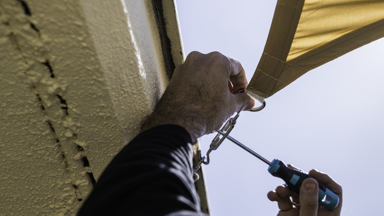 A man tightening a tensioner at the corner of a shade sail attached to the side of a home.